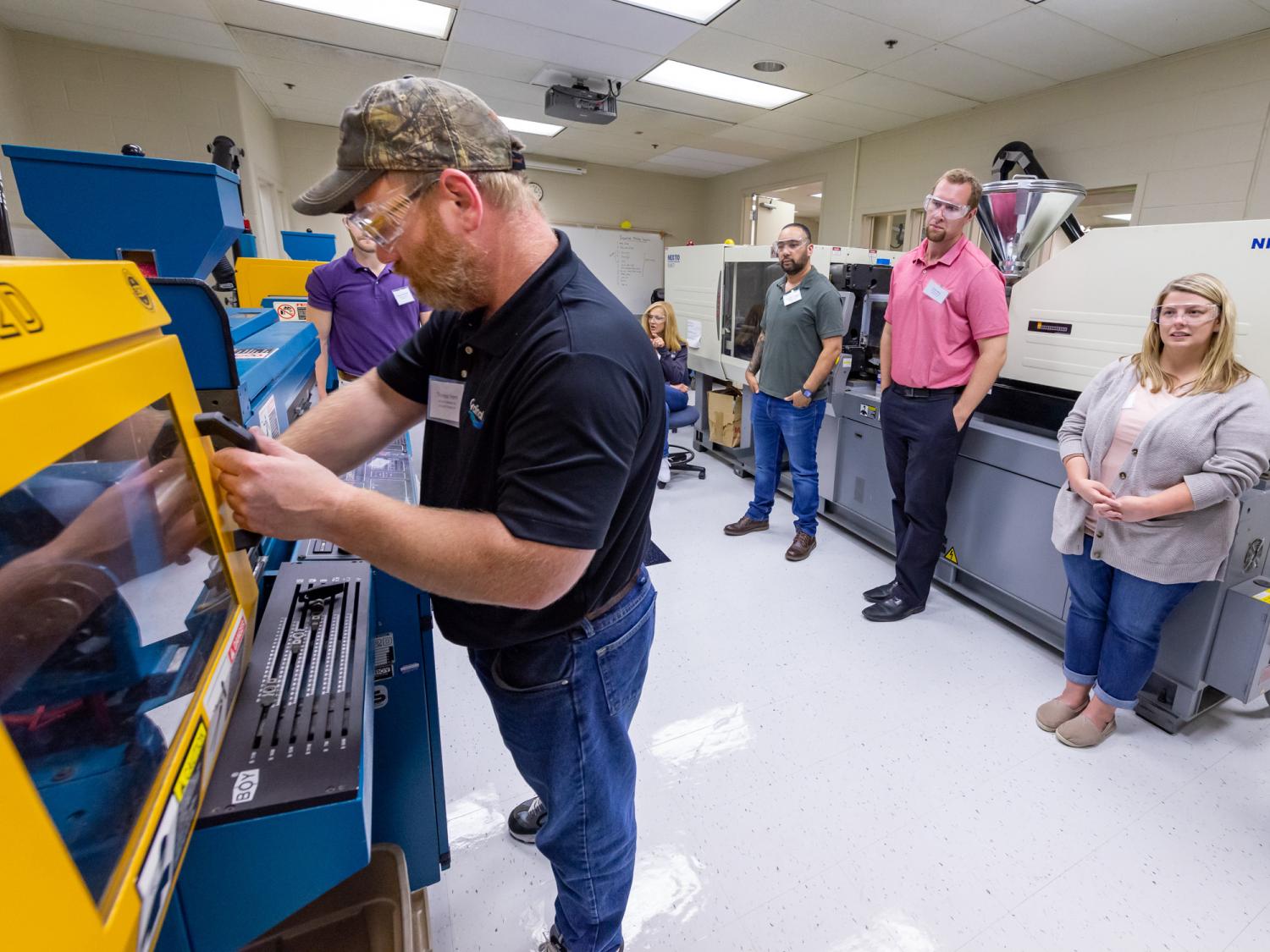 A man in a black shirt and blue jeans works in Penn College's injection molding lab while several other people watch.