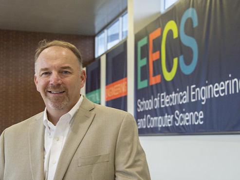 A man in a tan suit jacket and white button-down shirt smiles at the camera while standing in front of a large banner that reads "School of Electrical Engineering and Computer Science".