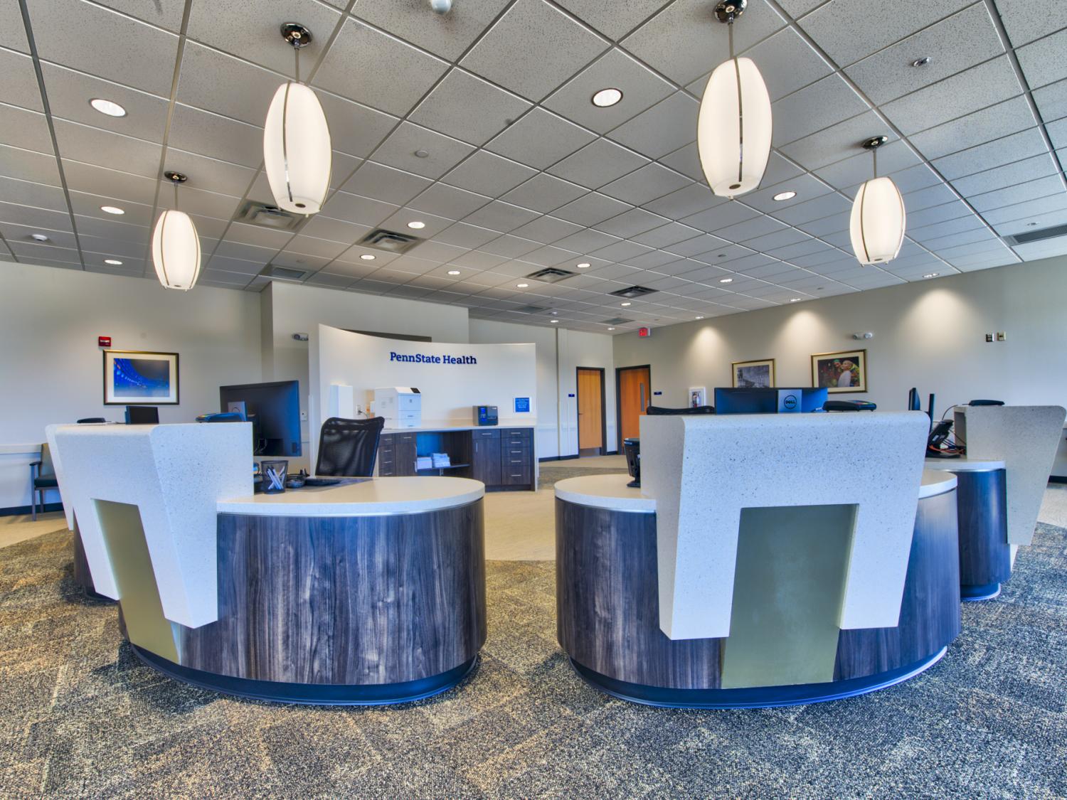 Two blue and white kiosks are in the foreground; a wall with the Penn State Health logo is in the background. Oval-shaped lights hang from the ceiling.