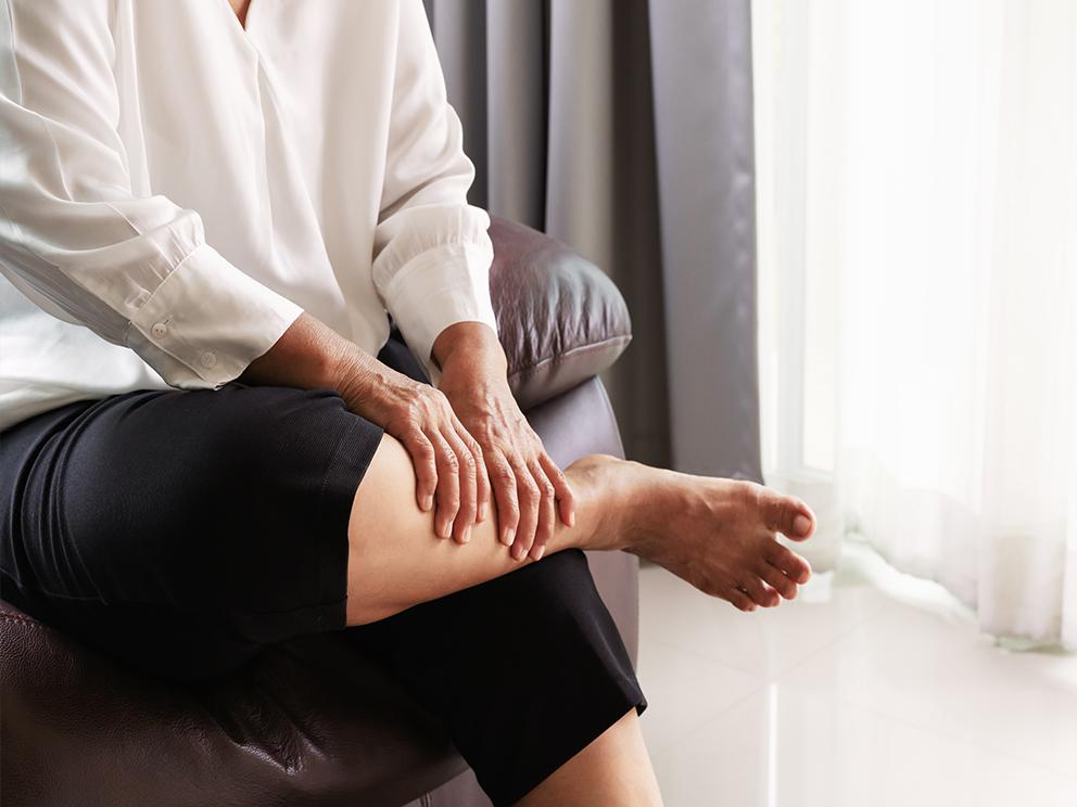 A woman sits on a couch, leaning forward. Her right leg is crossed over her left, and she holds the bottom part of her right leg with both hands.