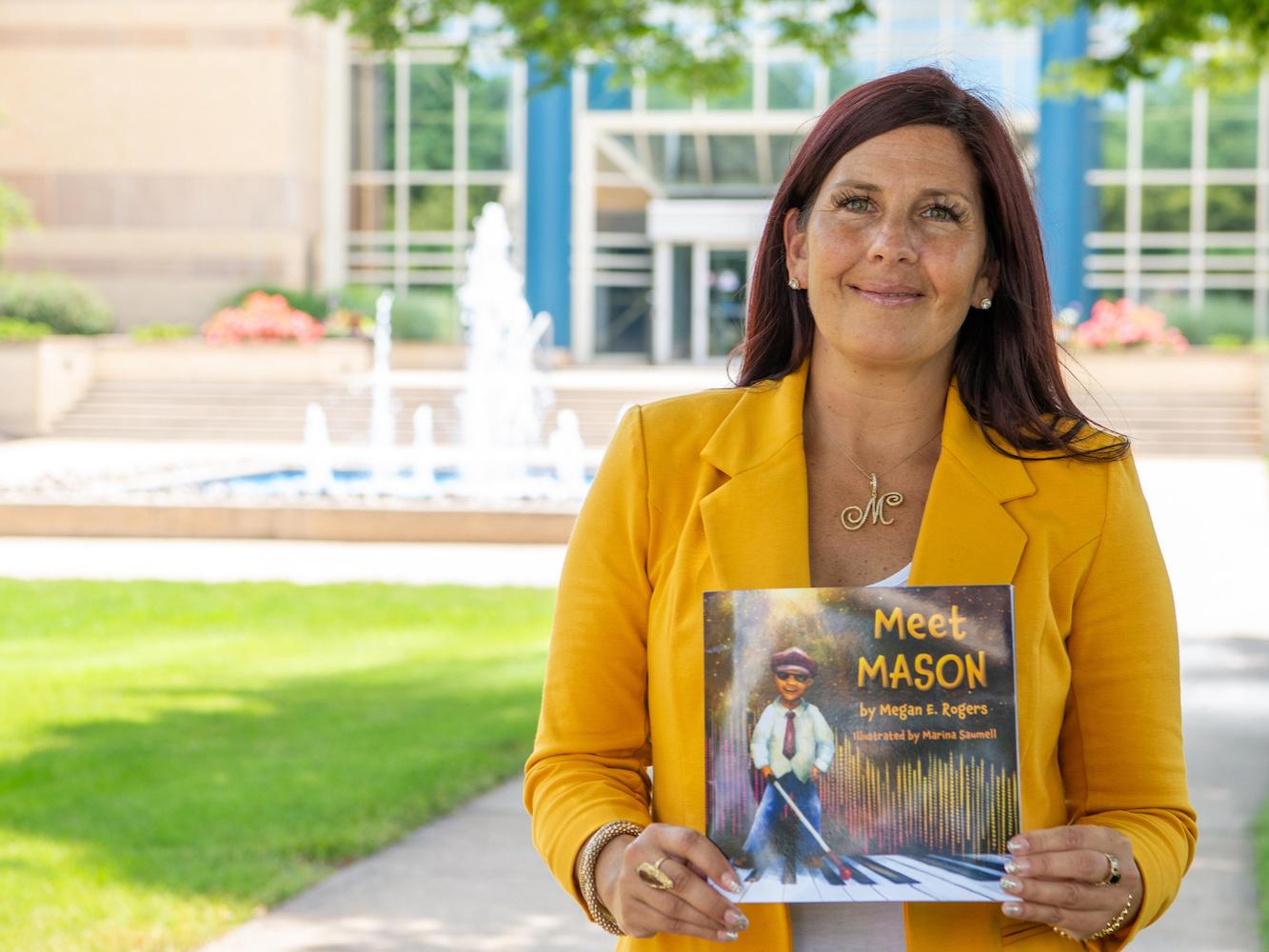 Megan E. Rogers stands wearing a yellow j while holding a copy of her book "Meet Mason"