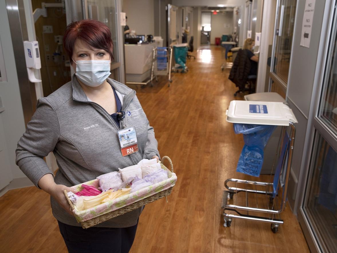A woman in a mask stands in a clinic hallway with stained-wood floors holding a basket of feminine hygiene products.