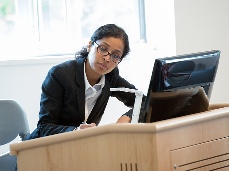 A faculty member stands at a lectern in front of a computer.