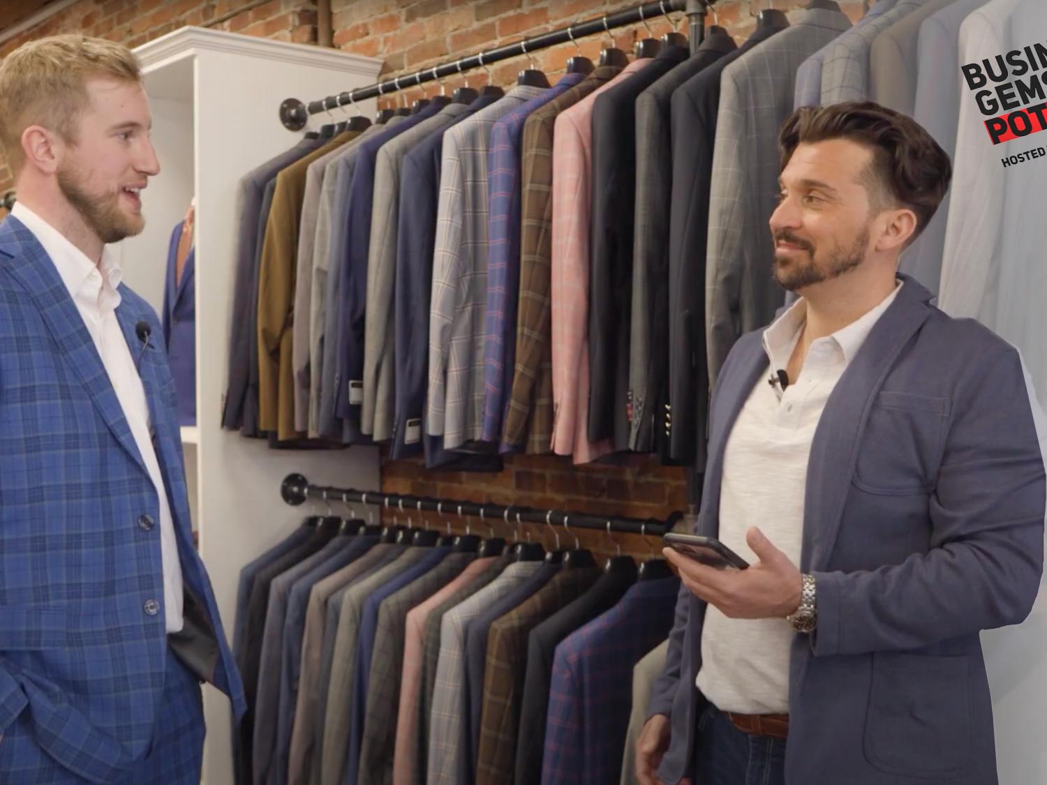 Two men wearing suits talk in a men's clothing boutique as they stand in front of sports jackets with the "Business Gems Pottsville" logo on the right