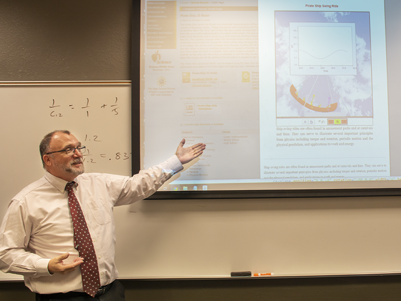 Michael Gallis stands in front of a white board with fractions written on it as he points to a projection screen showing the physics of an amusement park's pirate ship ride.