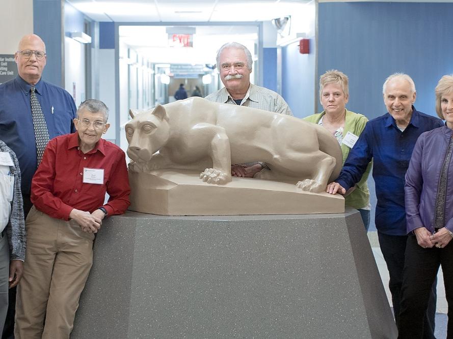 Nine participants from Penn State College of Medicine’s Mini Medical School pose in front of a Nittany Lion statue in the rotunda of Hershey Medical Center. Behind them a hallway is visible.