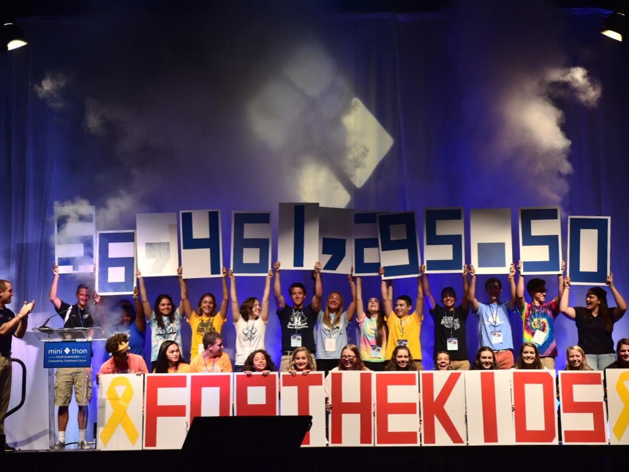 Several people stand on a stage and hold up numbers comprising the figure $6,461,295.50. In front of them are large red letters reading "FOR THE KIDS."