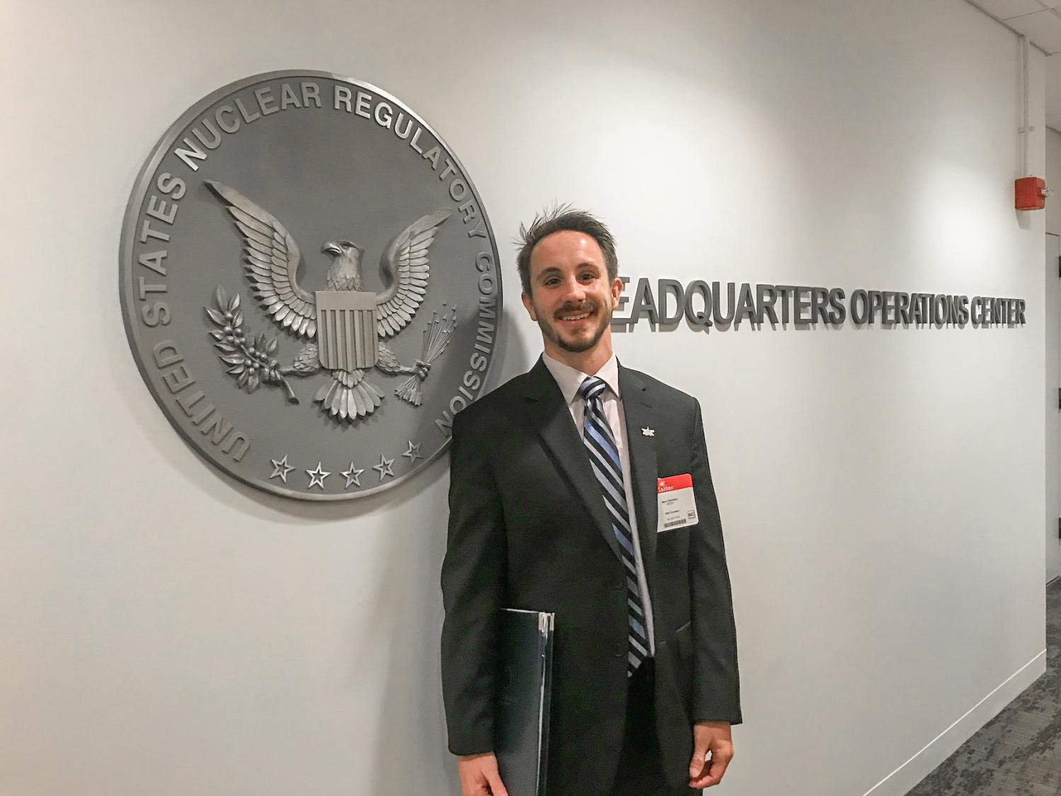 A man in a suit smiles and stands in front of a seal that reads "Unites States Nuclear Regulatory Commission."
