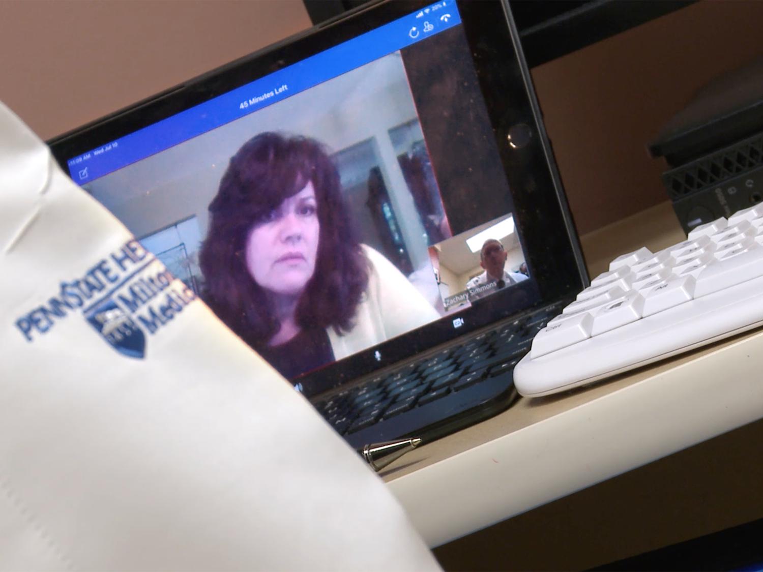 A close-up of Cynthia Lacey’s face is on a laptop monitor that is angled to the left. Dr. Simmons' arm with the Hershey Medical Center logo on it is visible in the foreground. Next to him is a keyboard.]