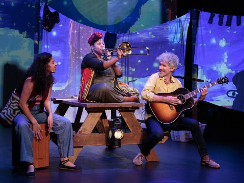 Three people sing and play instruments while sitting on a picnic table.