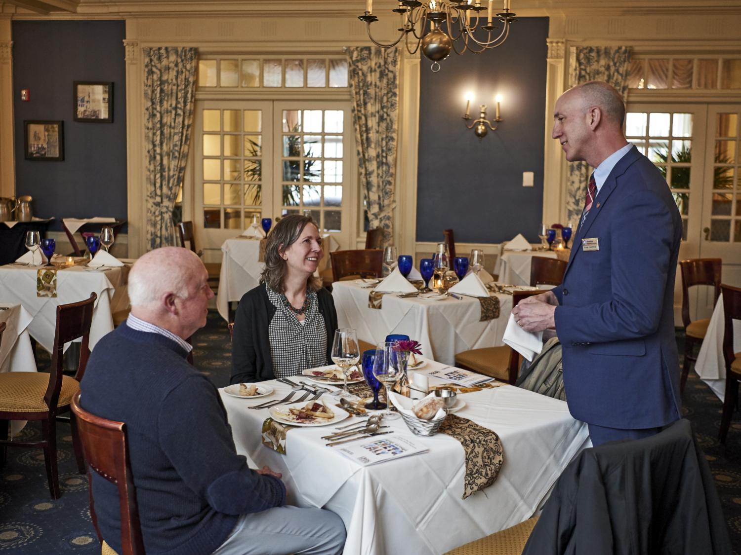 A man and a woman are seated at an elegant restaurant dining table, helped by a server.