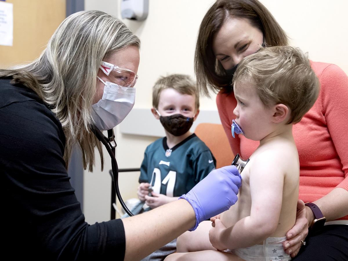 Dr. Melissa Tribuzio holds a stethoscope to Matthew Fitzpatrick’s chest. He wears a diaper and has a pacifier in his mouth. Samantha Fitzpatrick, who is wearing a sweater and a mask, places her hand behind his back. A young boy is in the background.