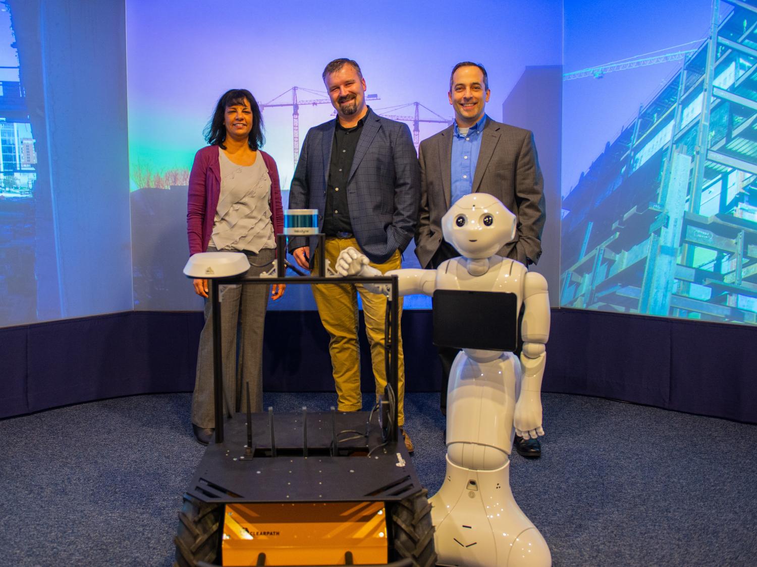 one woman and two men stand behind two medium-sized robots; a construction robot with wheels and a white robot shaped with human-like arms and eyes.
