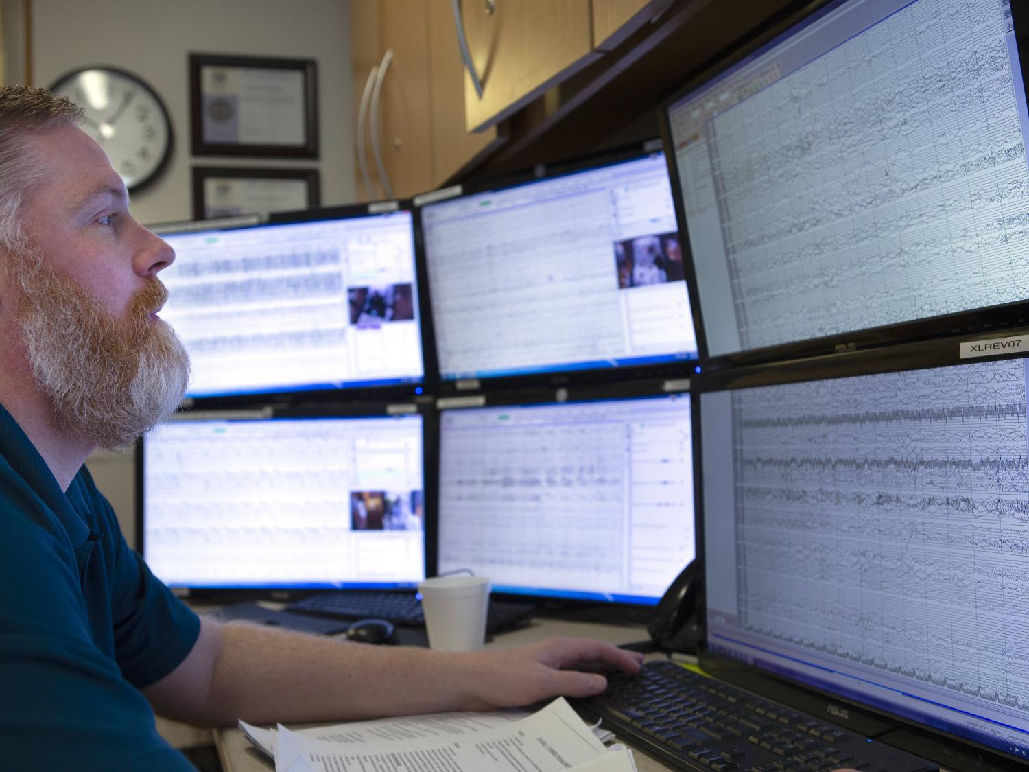 A man sits at a desk, at which six computer monitors, stacked in pairs, are in front of him.