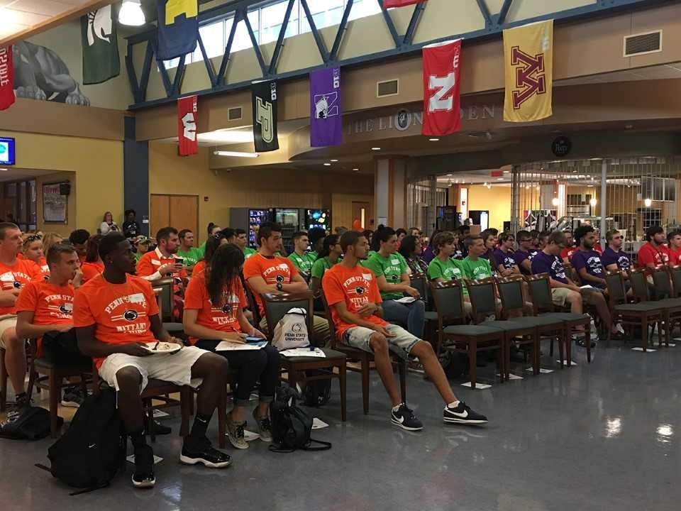 Approximately one hundred new students are seated in the campus's Great Hall wearing various colors of T-shirts.