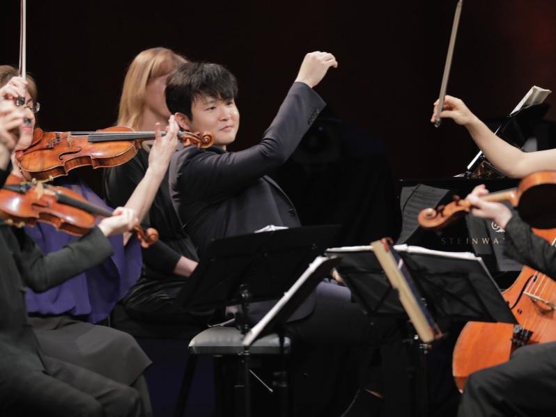 A musician plays his piano while violinists seated around him play their instruments.