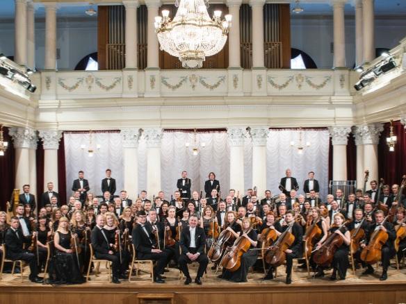 Musicians sit and pose with their instruments in an opulent hall.