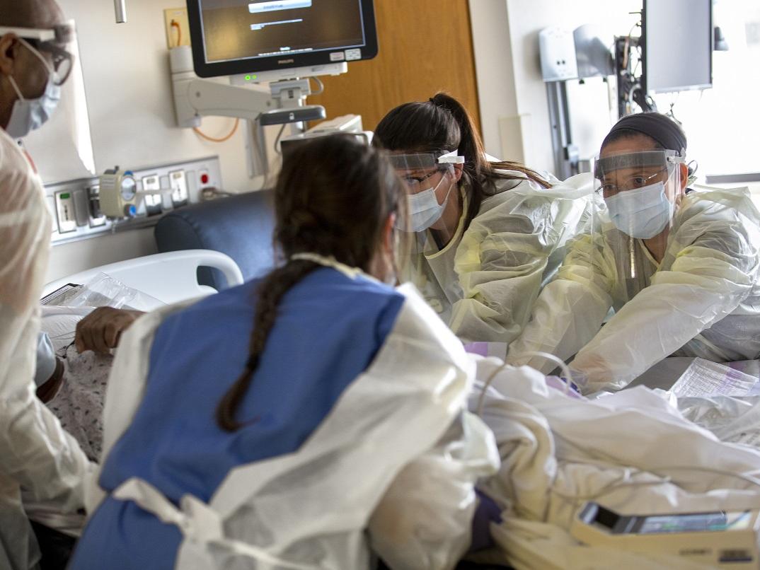 Two nurses, facing camera, with the Medical Intermediate Care Unit at Hershey Medical Center, and two other health care workers, whose backs are to the camera, touch a patient who is lying in bed. They are wearing gowns and face shields.
