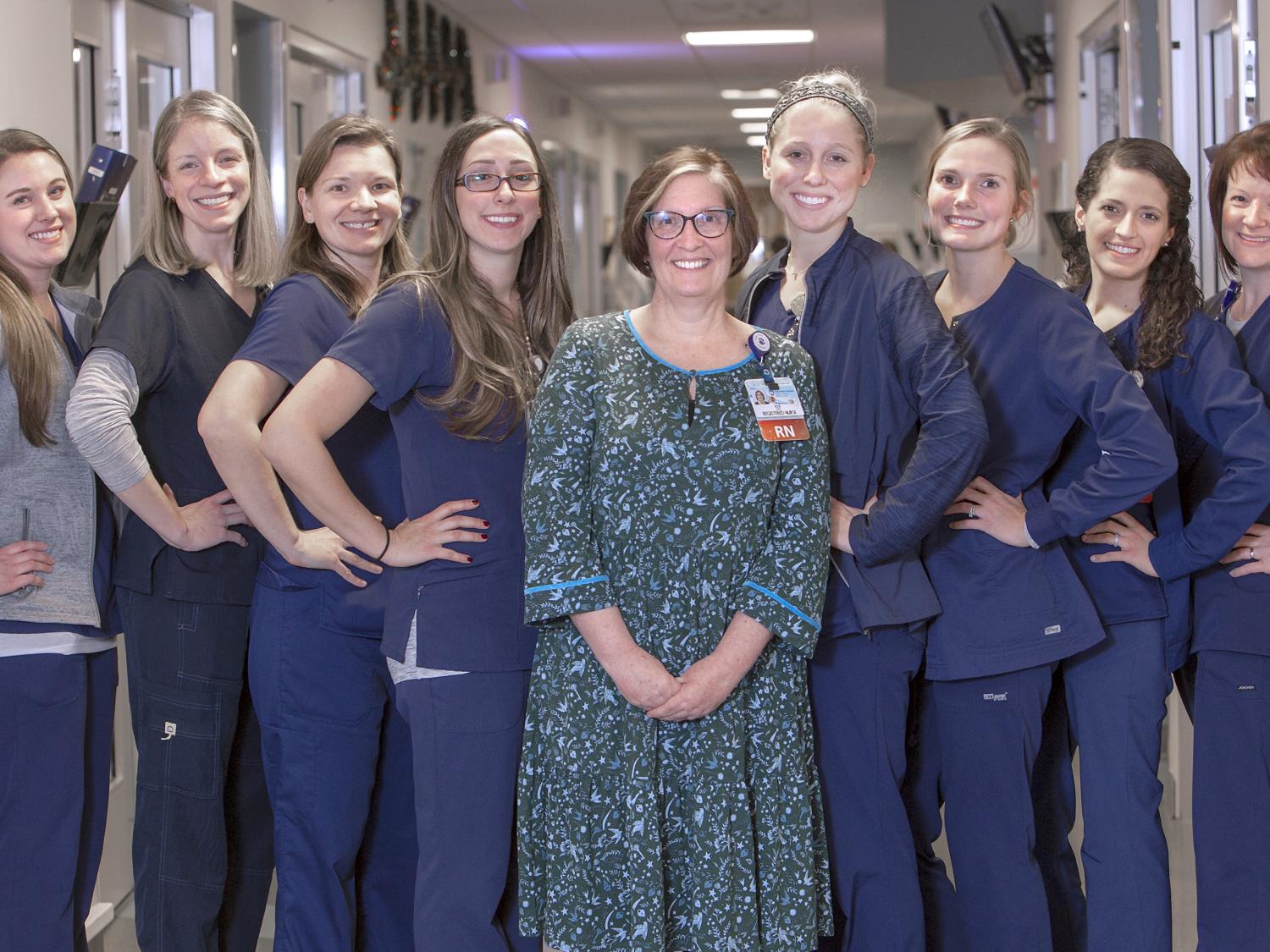 Nine women, all employees of the Heart and Vascular Progressive Care Unit at Hershey Medical Center, pose in the middle of the unit’s hallway. Julie Werner, nurse manager, stands in the middle of the group wearing a dress and ID badge. The other women stand on either side of her angled to the side with their hands on their hips smiling.