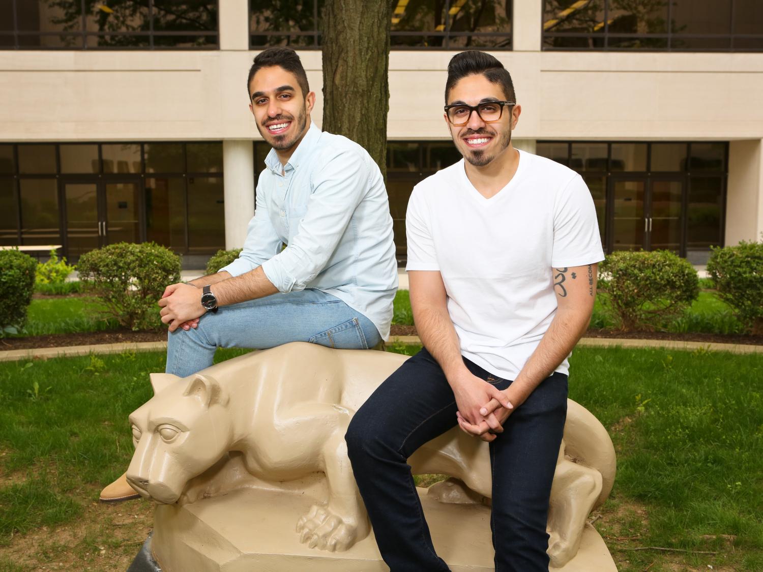 Two men sit on a Nittany Lion statue in the courtyard of Penn State College of Medicine. The building – beige with dark windows – is in the background.
