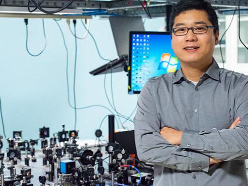 A man in a button-down shirt and glasses smiles at the camera in front of a computer and optical equipment.