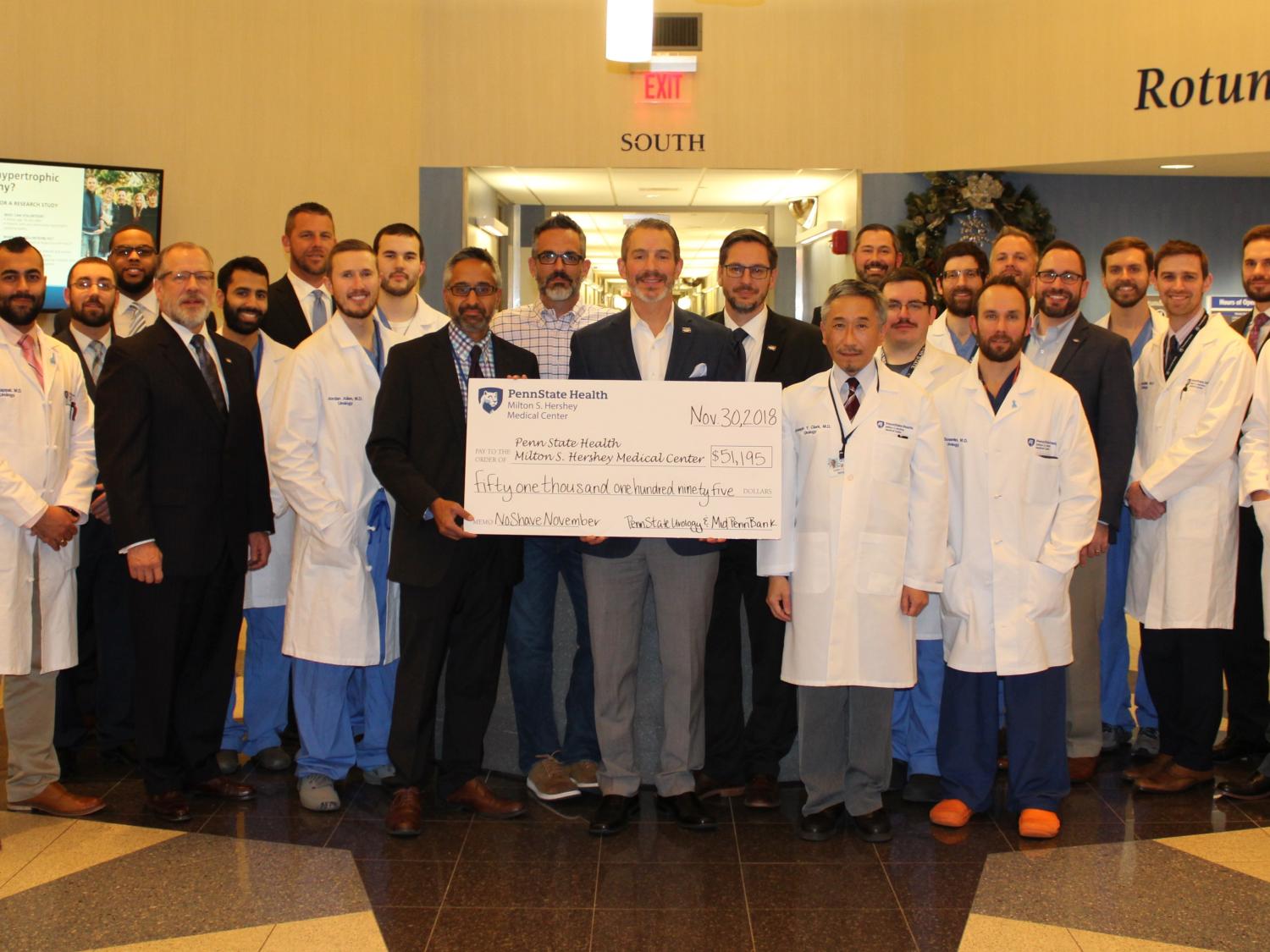 Dr. Jay Raman of the Division of Urology poses with Rory Ritrievi of Mid Penn Bank, surrounded by other No Shave November participants from both organizations. They pose holding an oversized check in the Rotunda at Penn State Health Milton S. Hershey Medical Center in brightly lit room with a TV screen behind them.