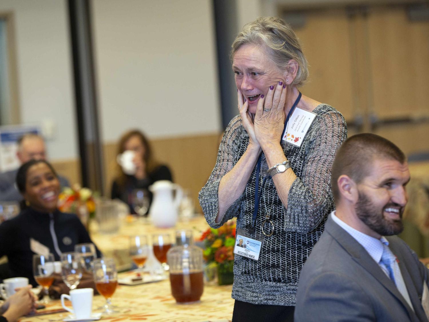 Judy Dillon’s hands cradle her smiling startled face. She stands at a table upon which are glasses and cups and saucers. Smiling people look on.