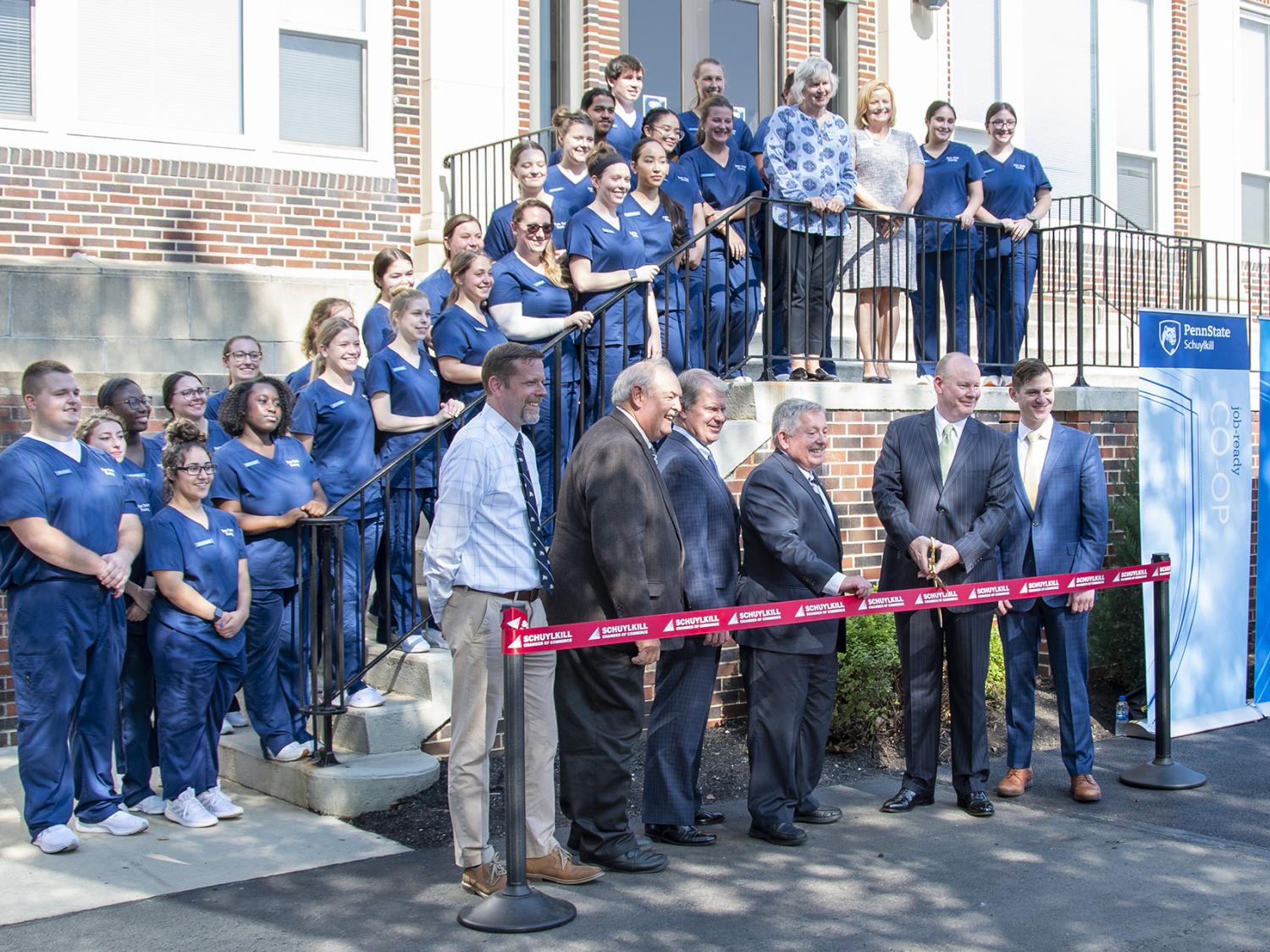 Students wearing dark blue scrubs line stairs in front of a brick building. Six men wearing suits stand in front of them while holding a red ribbon to be cut by large gold scissors.