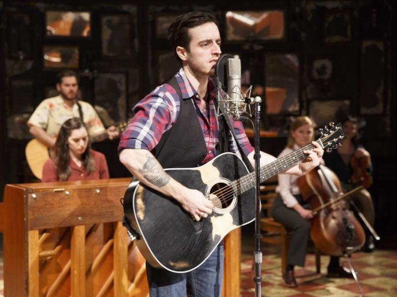 A male musician stands at a microphone and sings while he plays guitar.
