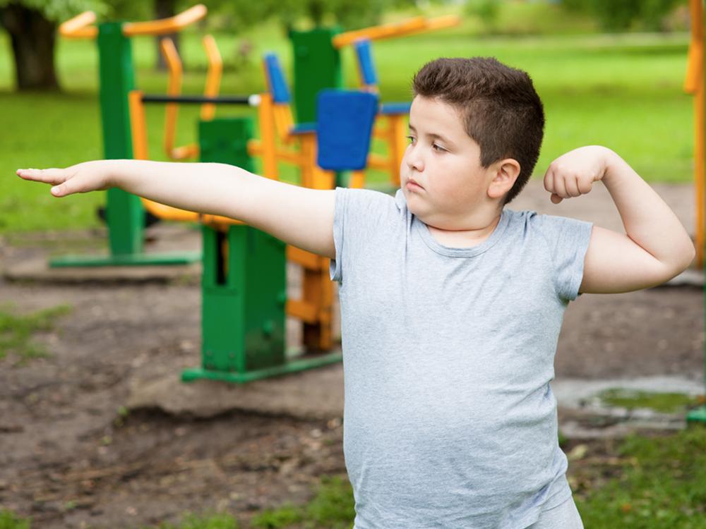 A young boy stands with his right arm extended outward and his left arm curled upward. Playground equipment is in the background.