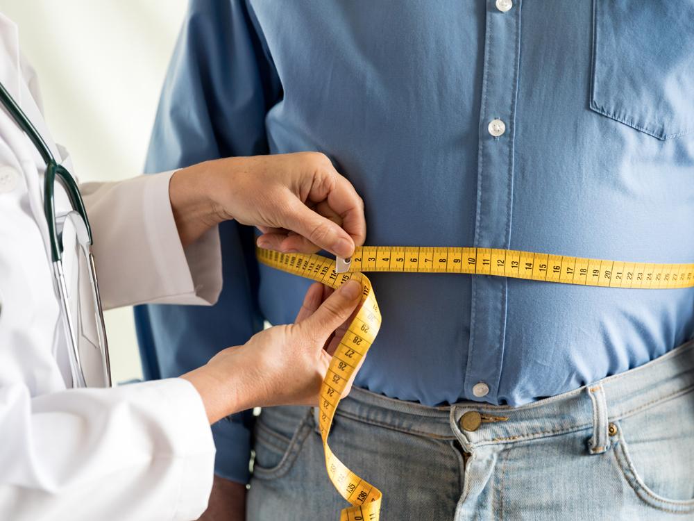 A physician in a white coat and a man in a blue button-down shirt and blue pants are seen from the torso down. The physician is holding a tape measure around the man’s waist.