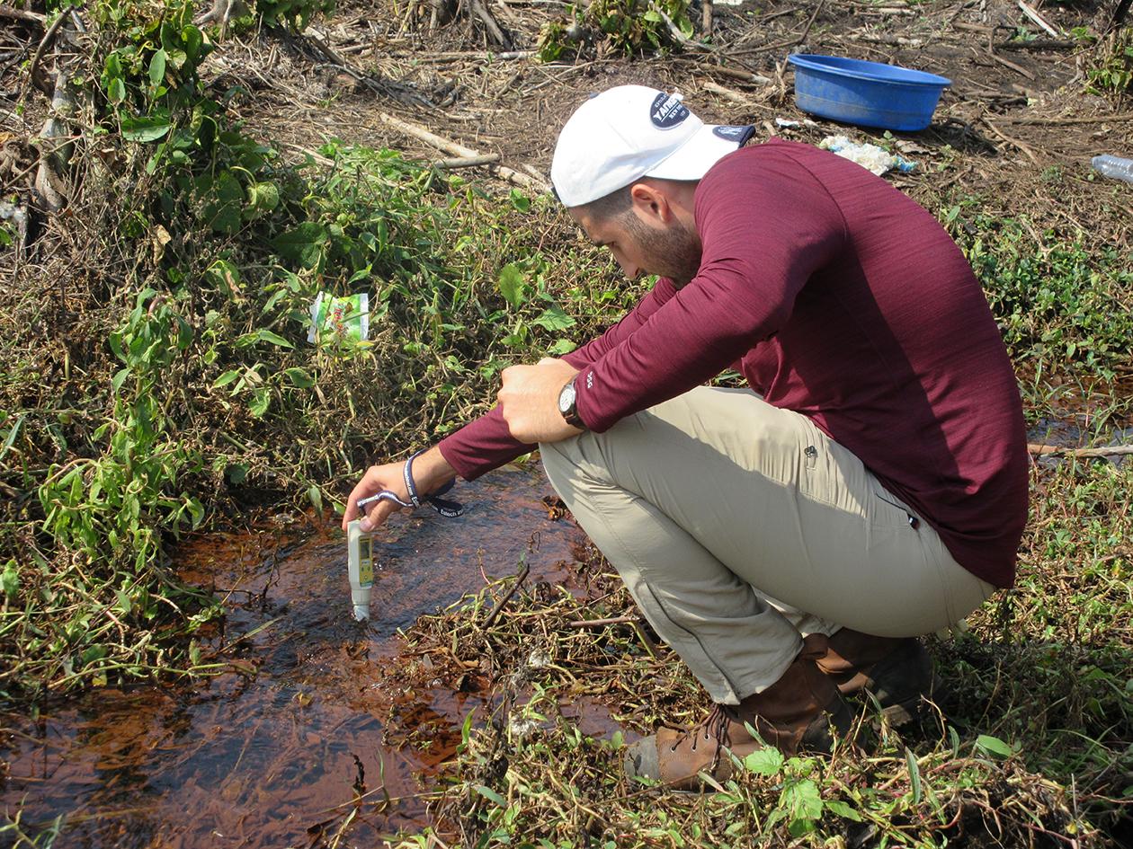 A man wearing a backwards baseball cap and a long-sleeve shirt and pants crouches in a marsh.