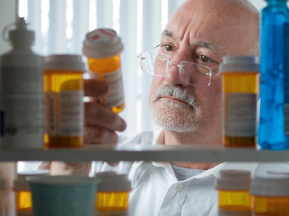 View from inside a medicine cabinet looking out, as a man in a white shirt holds an orange-brown pill bottle and looks at it. Other similar bottles are in the foreground.