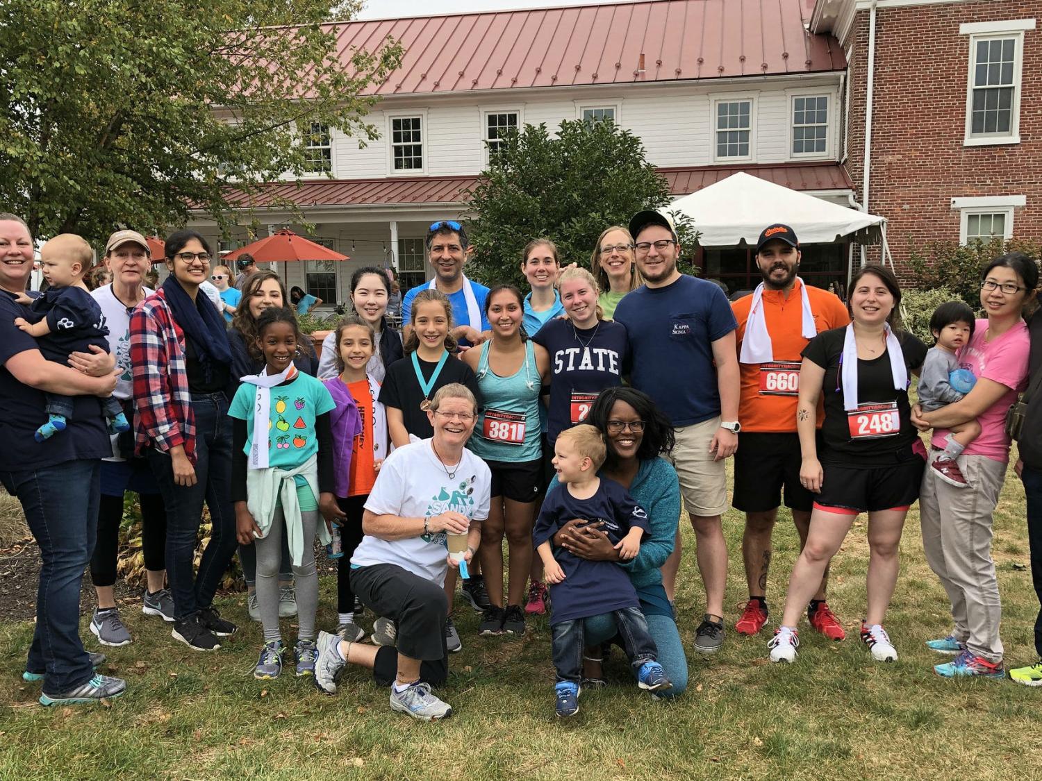 About a dozen participants from Penn State Health’s first annual Sandy Sprint pose on the lawn in front of a large brick house.
