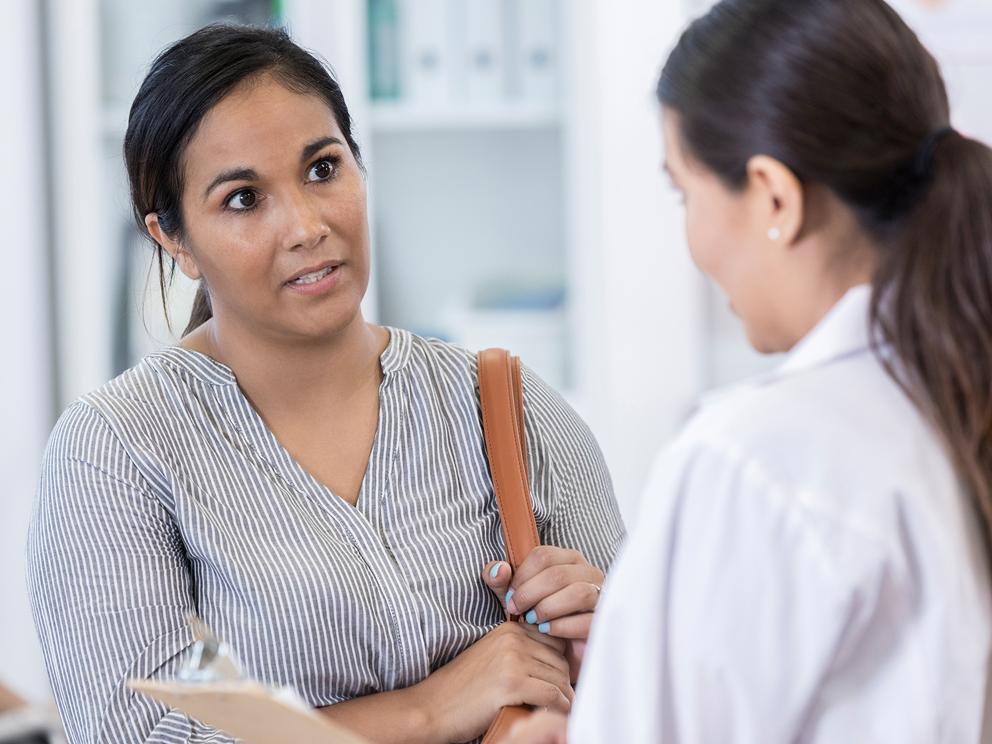 A woman in a striped shirt with a purse over her left shoulder speaks with a female doctor in a physician’s coat.