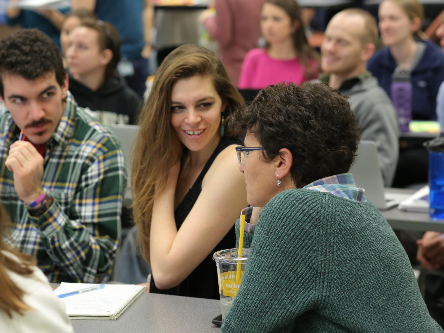 A room full of people sitting at tables. At the nearest table that’s in focus, a man and two women are talking.