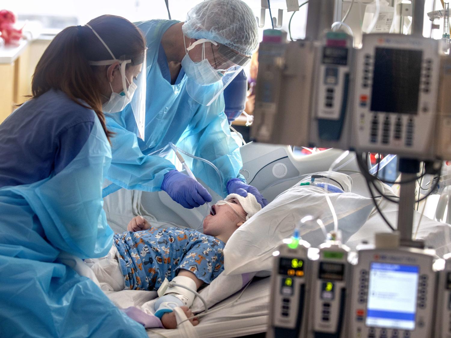 Two health care workers in personal protective equipment slide a tube down a boy’s throat, as the boy lies in a hospital bed.