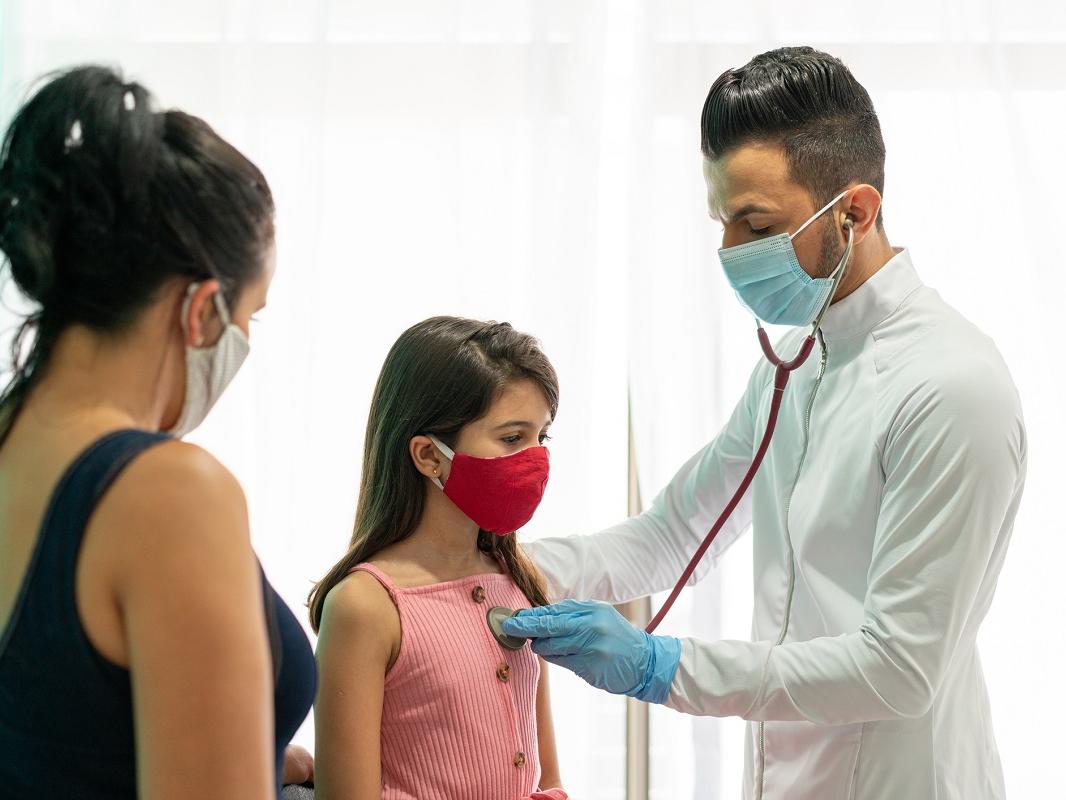 A doctor holds a stethoscope to the heart of a young girl as a woman looks on.