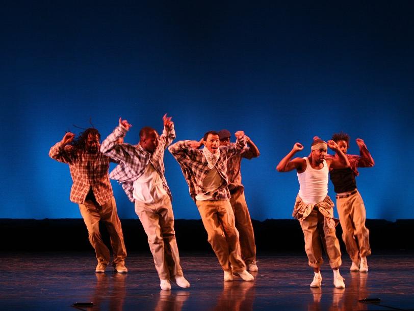 A small group of dancers in urban-style dress jump in unison while pumping their arms.