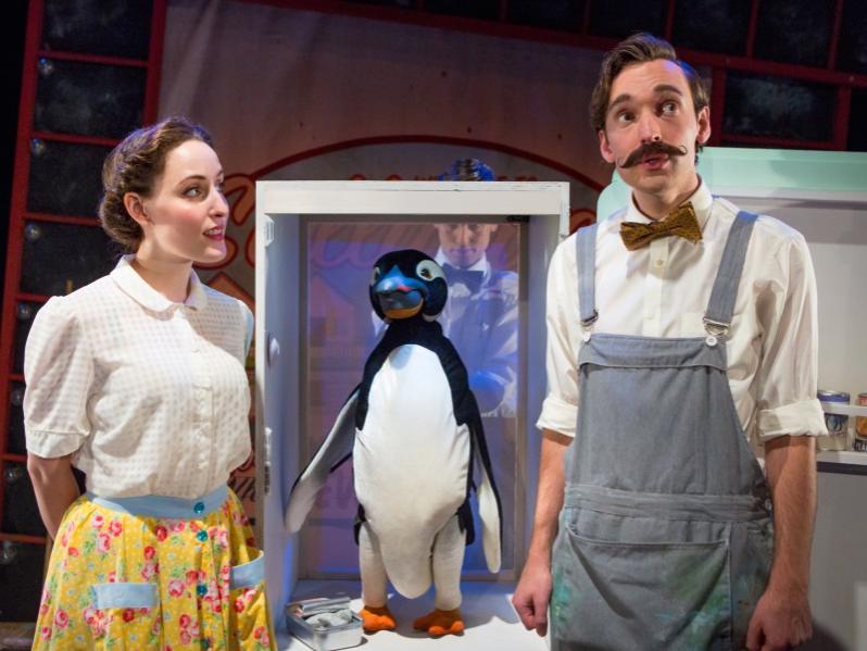 A woman and man dressed in early 20th-century clothing flank a penguin puppet perched on a stool.