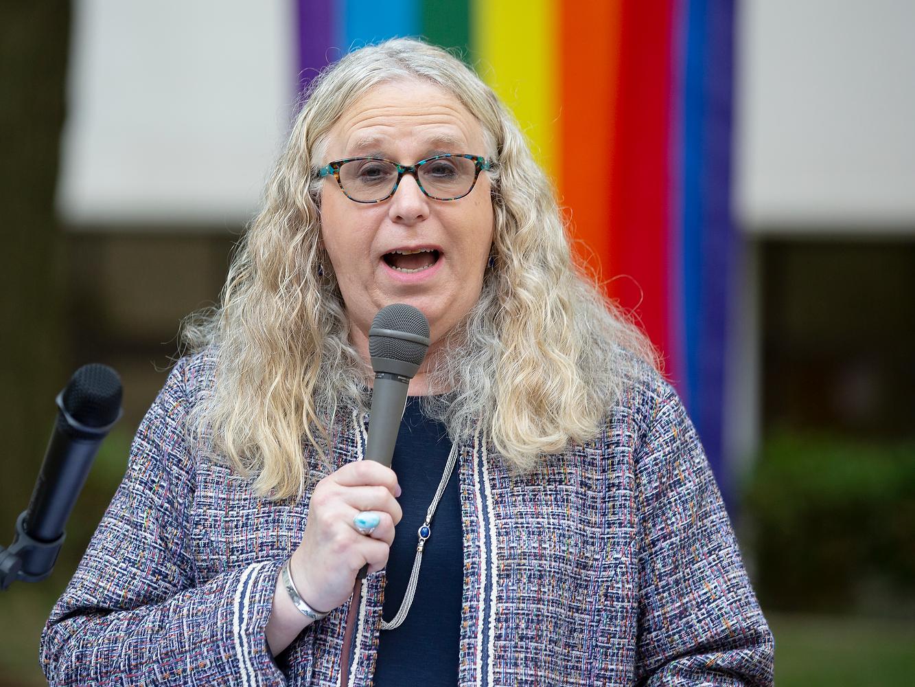 Rachel Levine speaks into a microphone. Behind her, a rainbow-colored flag is draped.