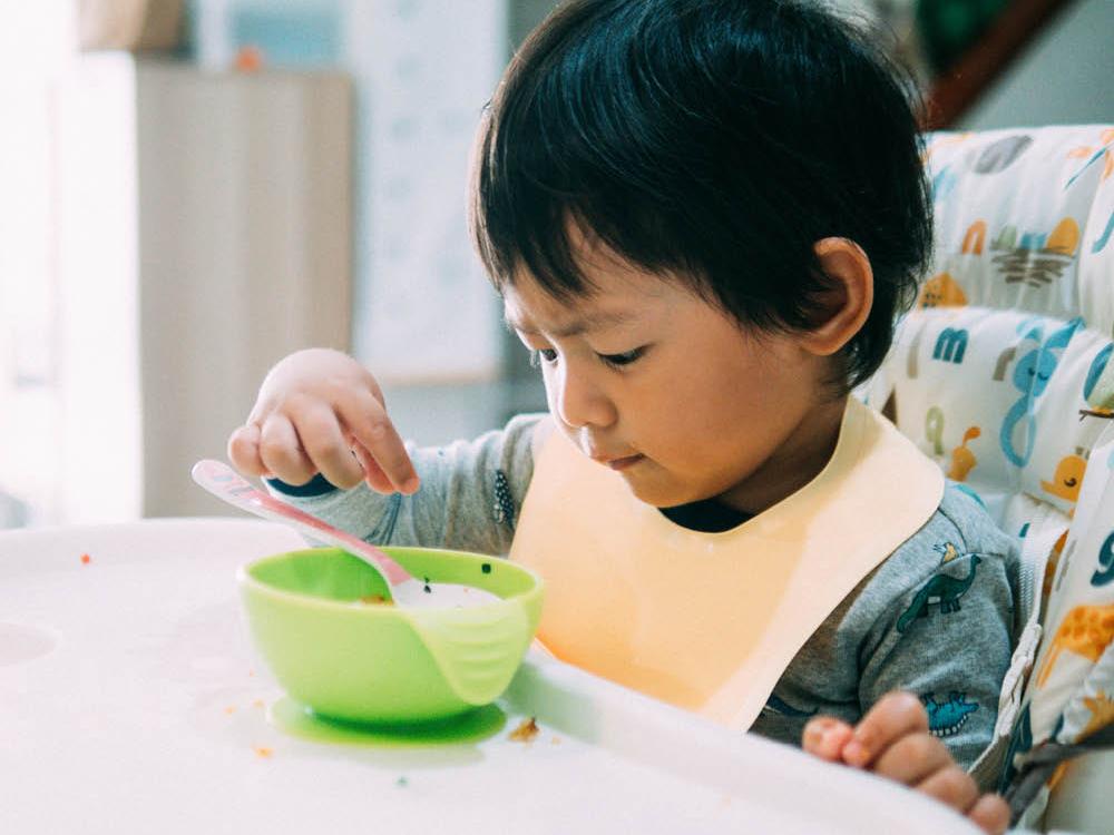 A young child sits at a table, a bowl and spoon in front of them
