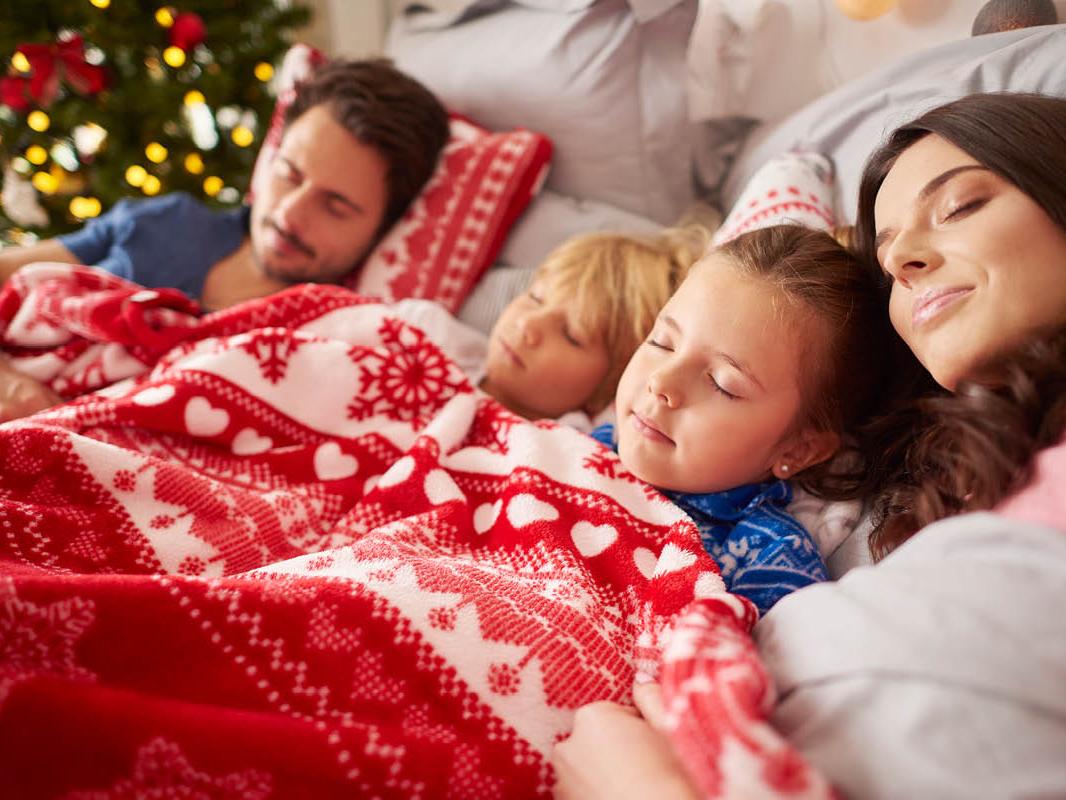 A man, woman and two young children lay down, sleeping, covered in a holiday-themed blanket. A lit Christmas tree is in the background.