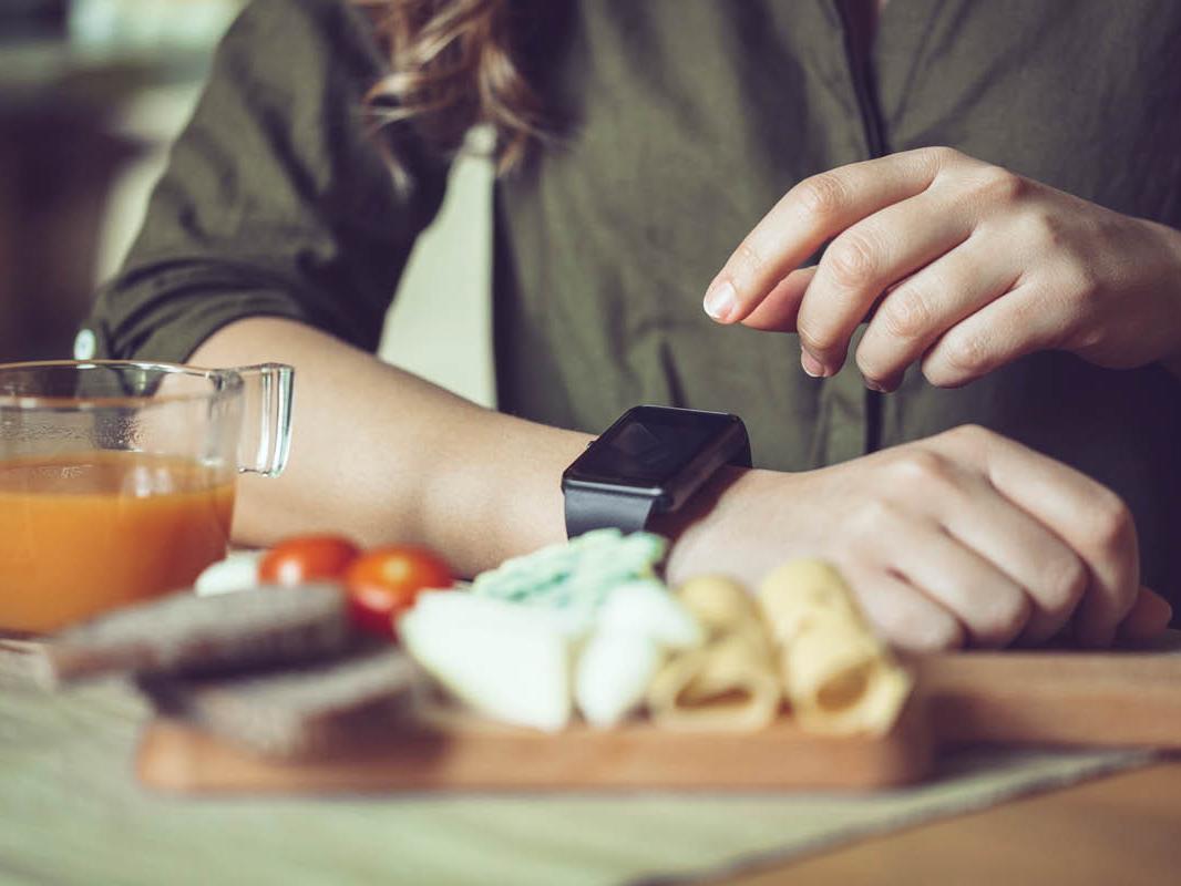 Close-up of a woman sitting at a table. She wears a square watch on her right arm, which rests on the table. Her left hand is reaching for the watch. Some food is on the table in the foreground.