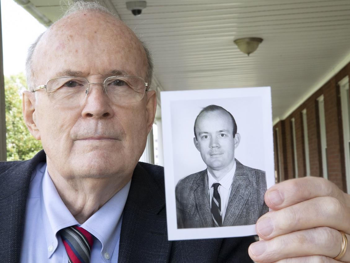 Dr. Cheston Berlin, who is wearing glasses and a suit and tie, shows a photo of himself from 1971.