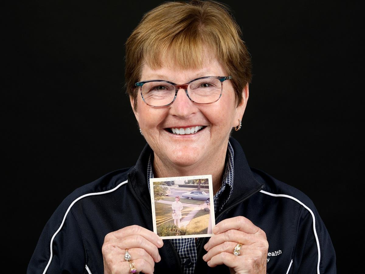 Janice Mills, who has short hair and glasses, smiles and holds a vintage picture of herself in a nurse’s uniform.