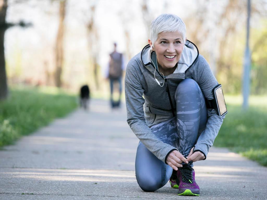 A woman in running tights and a jacket kneels to tie her left shoe. She is on a paved walking path bordered by grass, and a person and dog are in the background, out of focus. A smart phone is strapped to the woman’s left arm, and she has ear buds in.