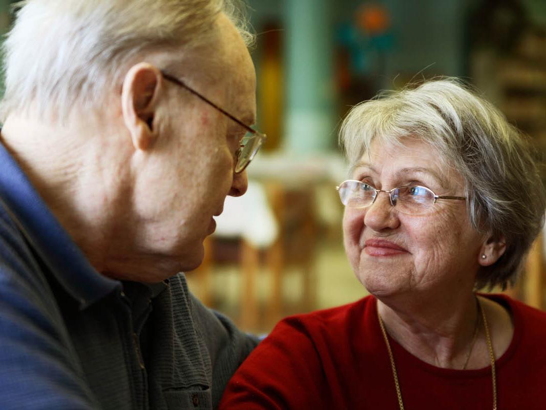 A man and a woman sit next to each other. The man has his arm around the woman. The two look at each other, smiling slightly.