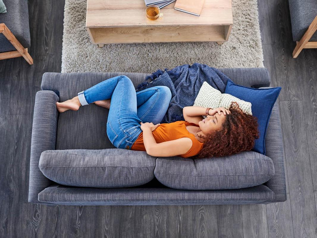 Overhead view of a woman with long hair laying on her right side, body slightly curled up, on a love seat. Her left hand is on her abdomen, her right hand is on her head, in apparent pain.