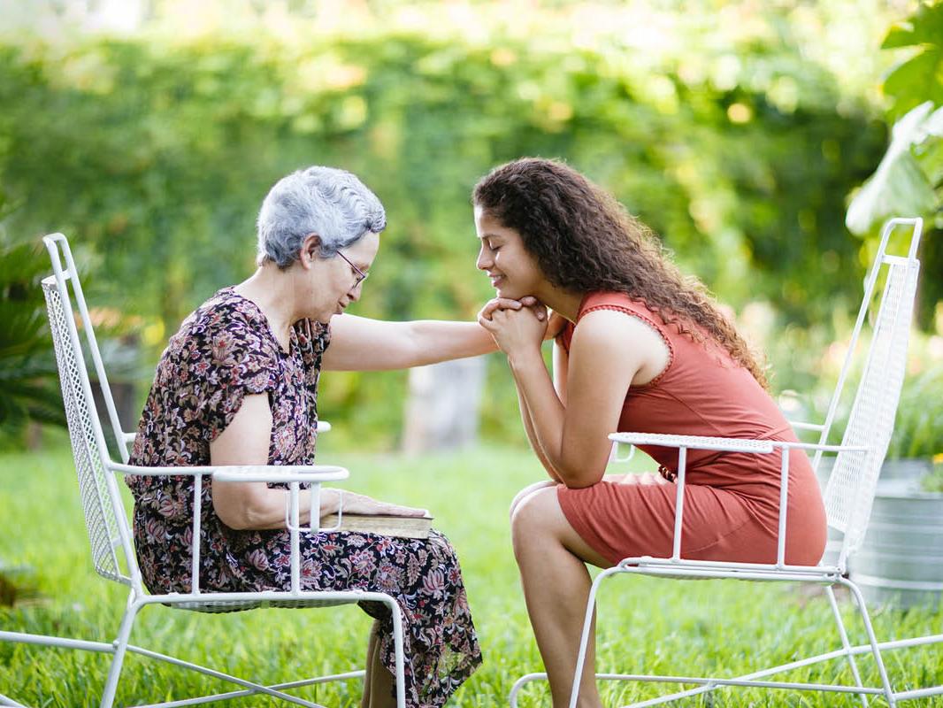 Two women sit in chairs, outdoors, facing each other. One woman has her left arm on the other’s right shoulder. Both lean forward slightly. Trees and grass are in the background.